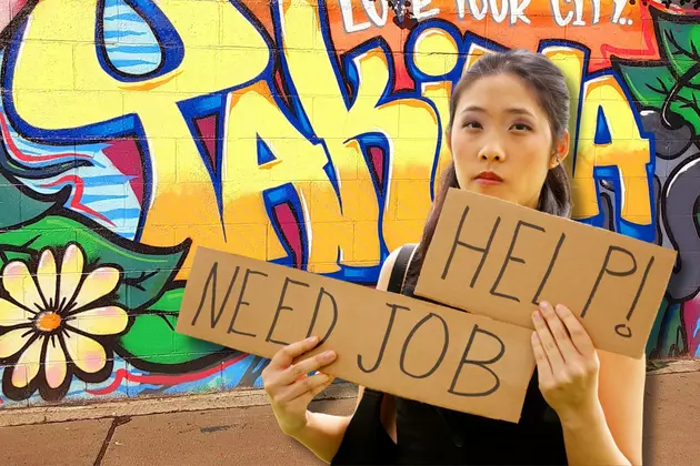 Woman holding a sign saying Help Need Job. Painted wall behind her saying Yakima